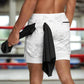 Man in a gym wearing light gray camo 2-in-1 gym shorts with a white inner compression layer, holding a black towel with another black towel tucked into the waistband, standing near a boxing ring.