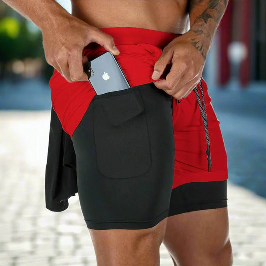 The image shows a close-up of a man wearing red 2-in-1 gym sports shorts with a black inner compression layer. The inner layer features a side pocket where the man is placing a smartphone. The red outer shorts have a drawstring waistband for an adjustable fit, and the black compression shorts underneath offer support and practicality during workouts. The outdoor background, with a blurred street scene, suggests that these shorts are versatile for both athletic and casual use, combining style and functionali