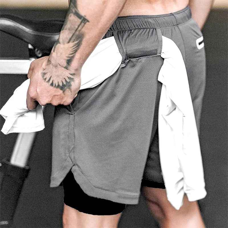 The image shows a close-up of a man wearing gray 2-in-1 gym sports shorts with a black inner compression layer. A white towel is tucked into the waistband at the back. The man's tattooed arm is visible as he holds another portion of the towel. The shorts feature an elastic waistband, providing a comfortable and secure fit during workouts. The background suggests an indoor gym setting, likely emphasizing the shorts' functionality and versatility for activities like weightlifting or cardio workouts.