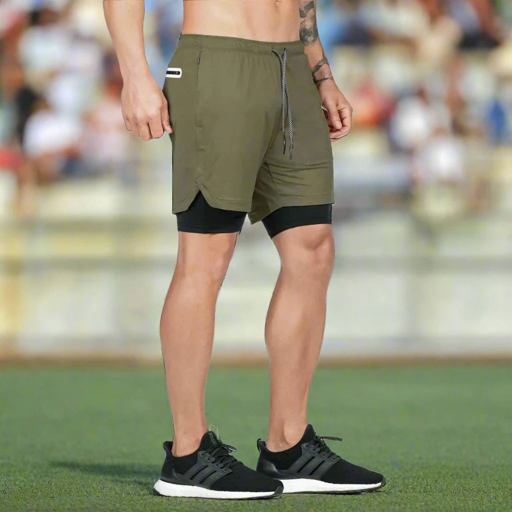 Man wearing olive green 2-in-1 gym shorts with a black inner compression layer, standing on a sports field in black athletic shoes.