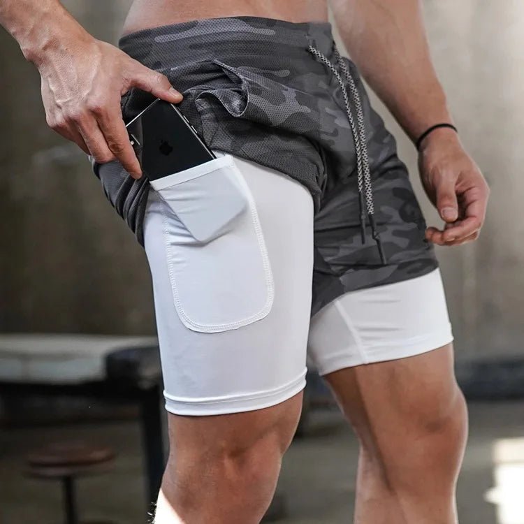 Man wearing gray camo 2-in-1 gym shorts, placing a phone in the side pocket of the white inner compression layer