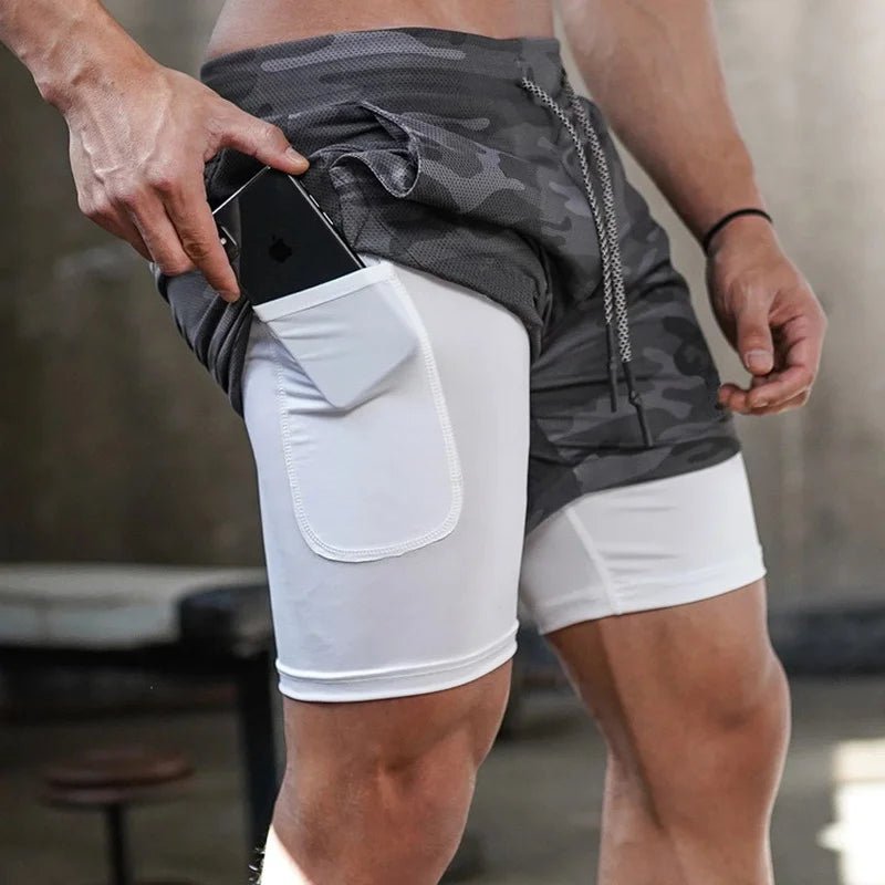 The image shows a close-up of a man wearing 2-in-1 gym sports shorts with a camouflage-patterned outer layer and a white compression inner layer. The inner layer features a side pocket, and the man is placing a smartphone into this pocket. The pocket is snug and designed to securely hold a phone or other small items during workouts. The shorts also have a drawstring waistband for adjustable comfort, making them functional and practical for athletic activities. The setting appears to be in a gym or workout s