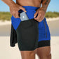 The image shows a close-up of a man wearing blue 2-in-1 gym sports shorts with a black inner compression layer. The inner compression layer features a side pocket where the man is placing a smartphone. The blue outer shorts have a drawstring waistband for an adjustable fit, while the black inner shorts provide support and practicality. The background appears to be an outdoor setting, possibly on a beach, highlighting the versatility of these shorts for both athletic and casual use in different environments.