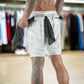 Man in a store wearing light camo 2-in-1 gym shorts with a black towel tucked into the waistband, holding another towel, showcasing detailed leg tattoos.