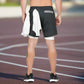 The image shows a man standing on a track, viewed from the back, wearing black athletic shorts with a white inner compression layer. A white towel is tucked into the waistband of the shorts. The shorts feature a zippered pocket near the back. The overall setting appears to be outdoors, on a running track, suggesting that the man is engaged in athletic activities like running or training. The image showcases the versatility and functionality of the shorts, highlighting their use in active scenarios