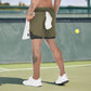 The image shows a man standing on a tennis court, viewed from the back, wearing olive-green 2-in-1 sports shorts with a black inner compression layer. A white towel is tucked into the waistband at the back, and he holds another white towel in his left hand. The man is wearing white athletic shoes, which complement the sporty setting of the tennis court. His leg tattoo is visible, and the shorts feature a minimalist design, combining functionality and style for active sports like tennis. The overall impressi