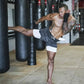 The image shows a man practicing martial arts or kickboxing in a gym, wearing 2-in-1 sports shorts with a black camouflage outer layer and a white inner compression layer. He is performing a high kick aimed at a heavy punching bag. His athletic build and tattoos are visible, and he is barefoot, suggesting a high-intensity training environment. The gym background features multiple punching bags, emphasizing a setting dedicated to martial arts or combat sports. The shorts are designed to provide flexibility, 