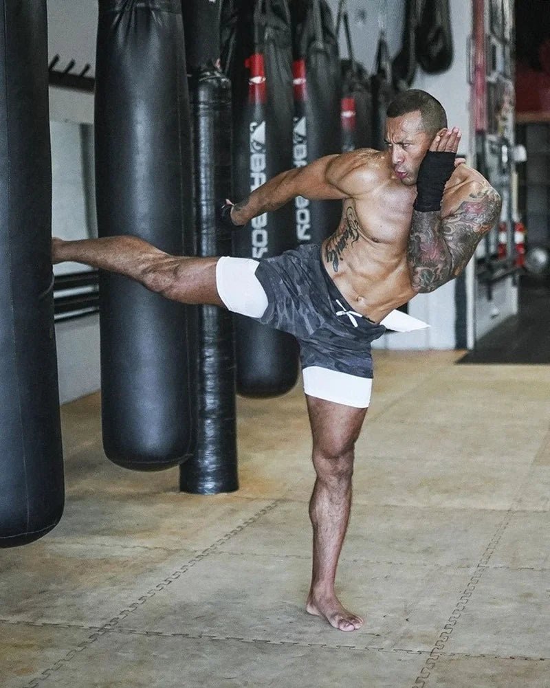 The image shows a man practicing martial arts or kickboxing in a gym, wearing 2-in-1 sports shorts with a black camouflage outer layer and a white inner compression layer. He is performing a high kick aimed at a heavy punching bag. His athletic build and tattoos are visible, and he is barefoot, suggesting a high-intensity training environment. The gym background features multiple punching bags, emphasizing a setting dedicated to martial arts or combat sports. The shorts are designed to provide flexibility, 