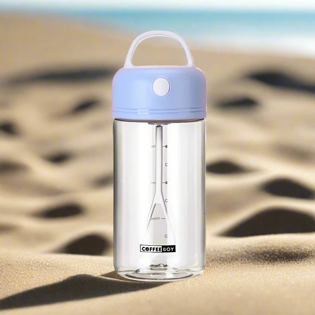 Electric shake bottle with a blue lid labeled 'Coffee Boy,' placed on sandy terrain with the ocean in the background.