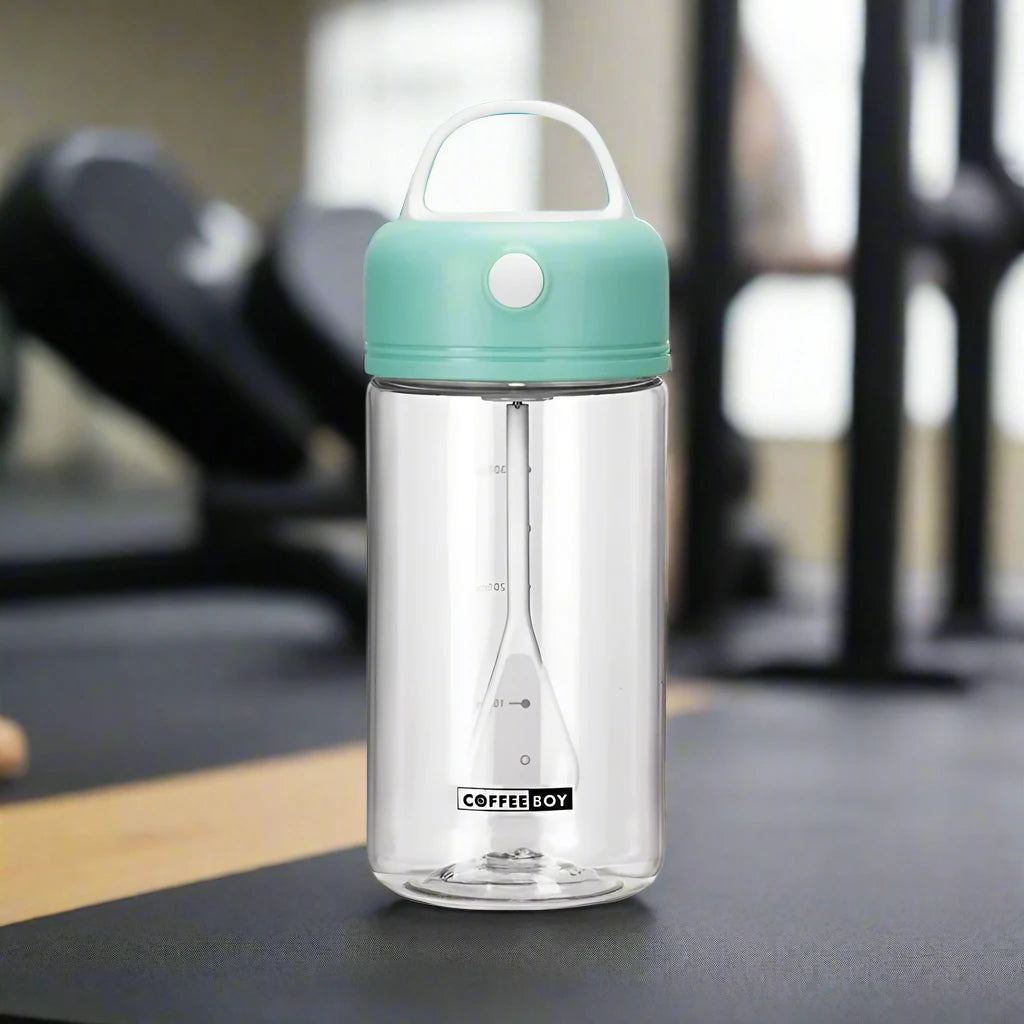 Electric shake bottle with a mint green lid labeled 'Coffee Boy,' placed on a gym bench with exercise equipment in the background