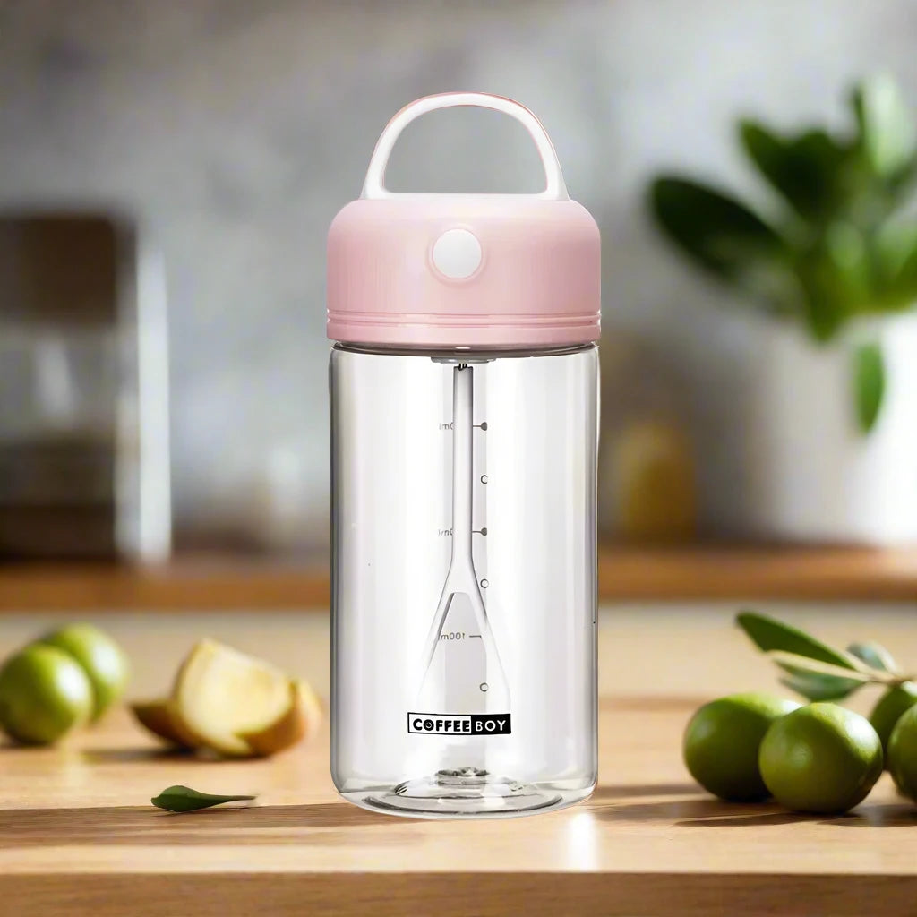 Electric shake bottle with a pink lid labeled 'Coffee Boy,' placed on a kitchen counter, surrounded by fruits and a plant.