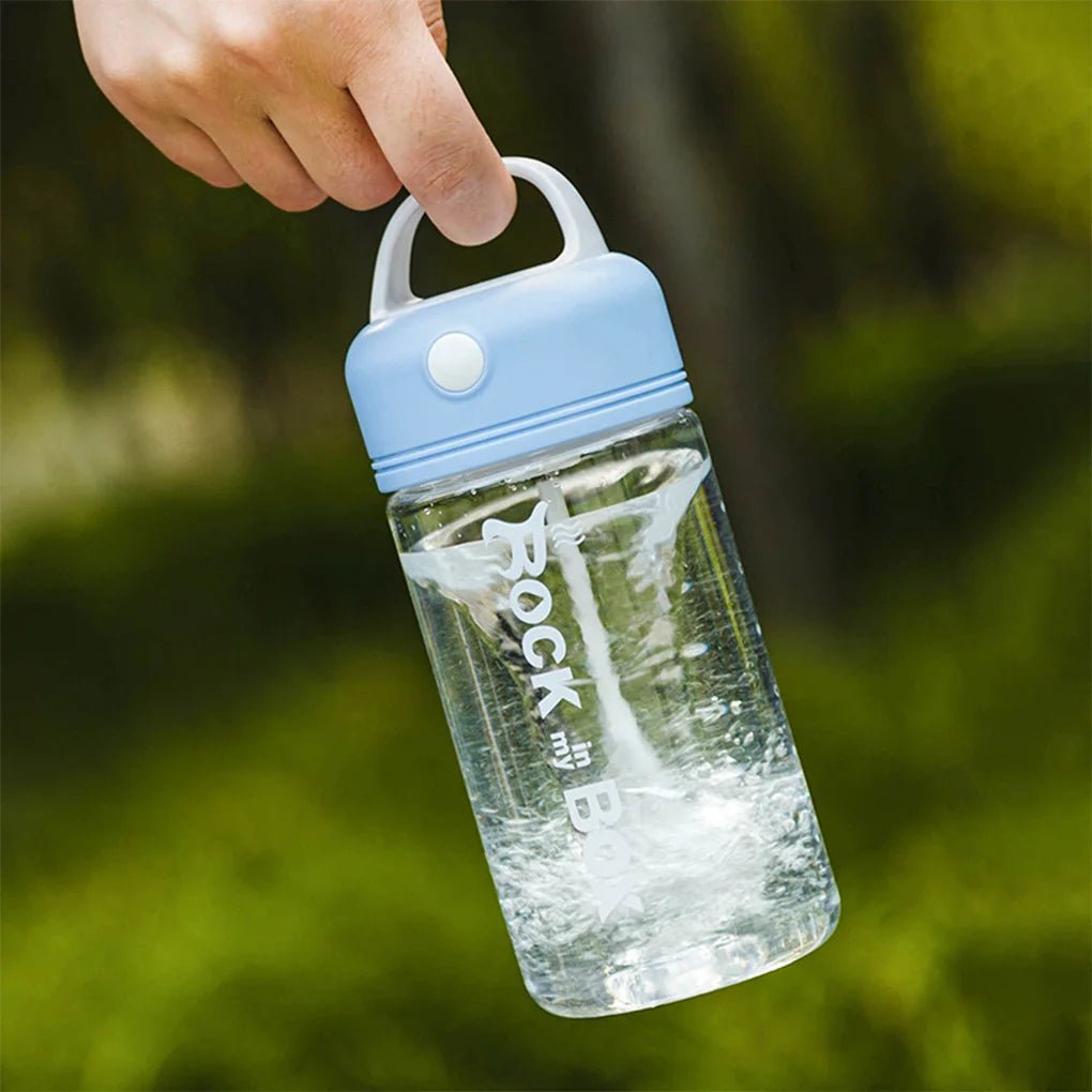 Hand holding a blue-lidded electric shake bottle labeled 'Rock in my Box,' with liquid swirling inside, outdoors against a green, blurred background.