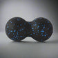 Black double massage ball with blue speckles, used for muscle relaxation and deep tissue therapy, placed on a gradient grey background.