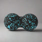 Black double massage ball with turquoise speckles, designed for deep tissue muscle relaxation, resting on a neutral grey background.