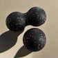 Black double massage ball with pink speckles, designed for muscle relaxation and therapy, placed on a light background.