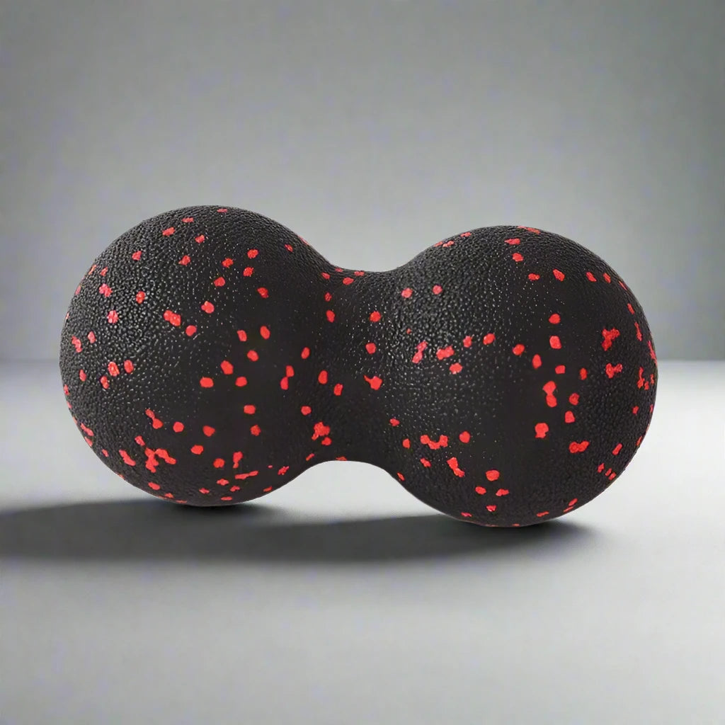 Black double massage ball with red speckles, designed for muscle relaxation and recovery, resting on a smooth grey surface.