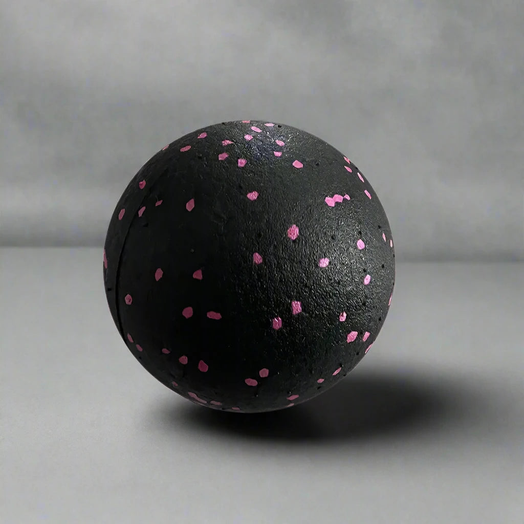 Black massage ball with pink speckles, designed for muscle therapy and tension relief, placed on a smooth grey surface.