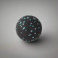 Single black massage ball with blue speckles, ideal for muscle tension relief, placed on a smooth grey surface.