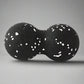 Black double massage ball with white speckles, designed for muscle therapy and tension relief, placed on a smooth grey surface.