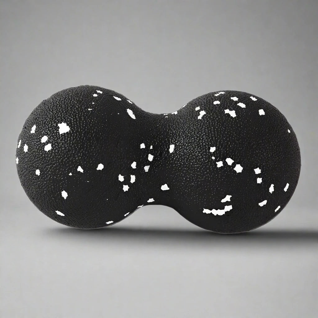 Black double massage ball with white speckles, designed for muscle therapy and tension relief, placed on a smooth grey surface.