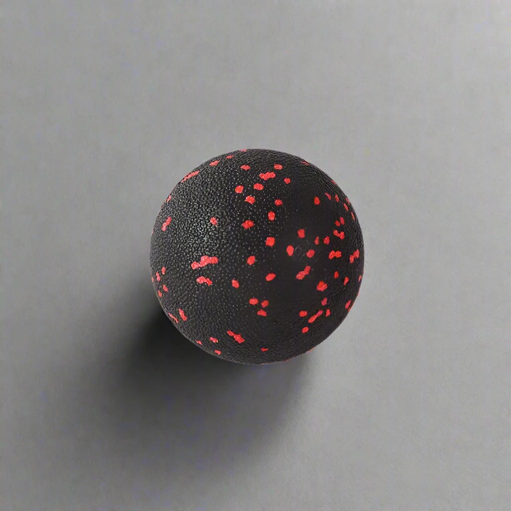 Single black massage ball with red speckles, designed for muscle relaxation and recovery, placed on a smooth grey surface.