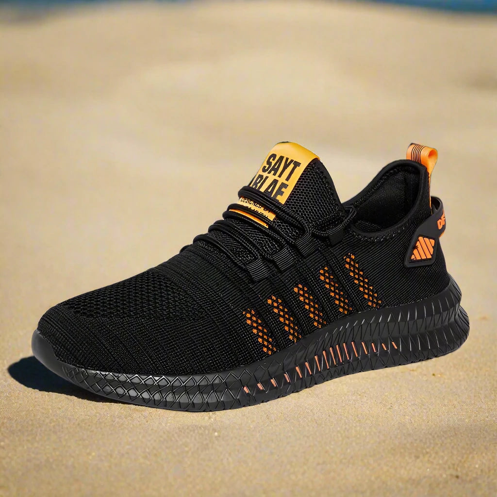 Black lightweight running shoe with orange accents, mesh upper, and textured sole for durability. Showcased on a sandy beach background.