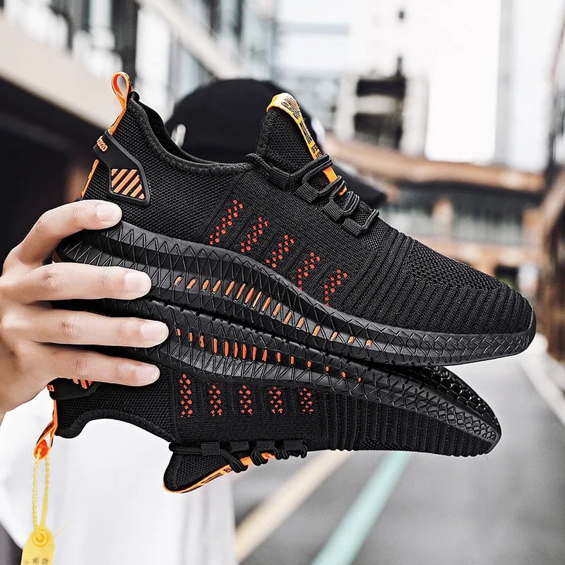 A close-up shot of a pair of black lightweight running shoes with orange accents and a textured sole, being held by a person in an urban setting. The sneakers feature a breathable mesh upper and a modern, streamlined design.